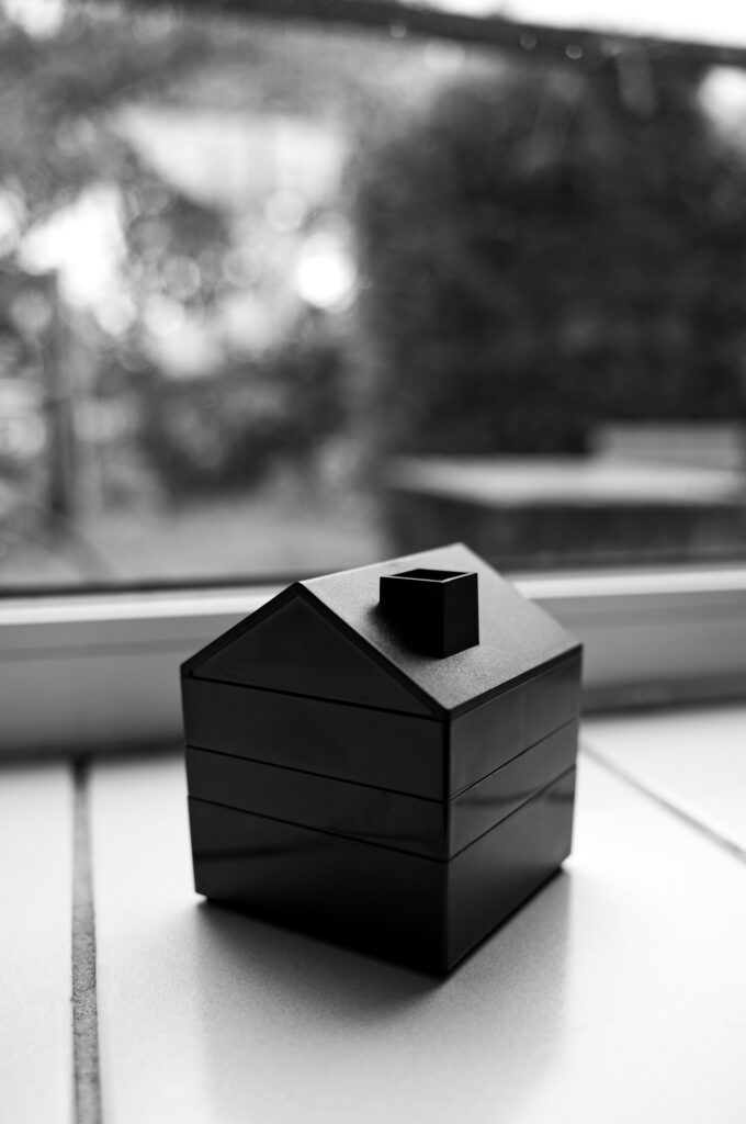 Photo of a small wooden or platic toy house in front of a window sill.